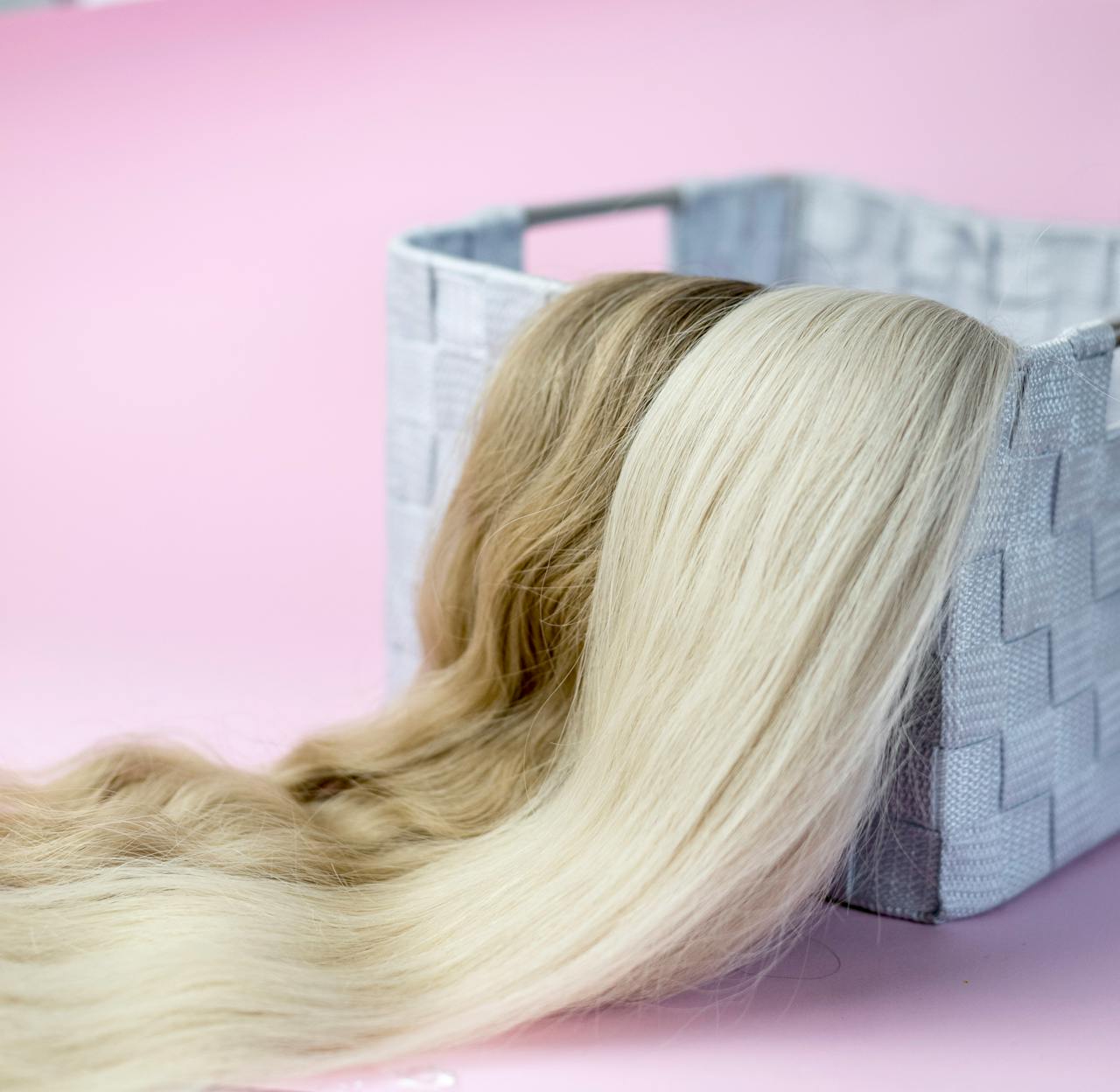 Close-up of blond and brown hair extensions draped over a woven basket, on a pink background.